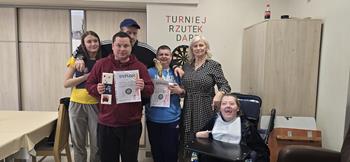 Uczestnicy trzymający puchar oraz dyplomy, oraz kadra ŚDS na tle napisu „turniej rzutek dart”